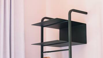 Un estante metálico negro con un perchero de madera, fijado a la pared, con una cortina a medio ver.