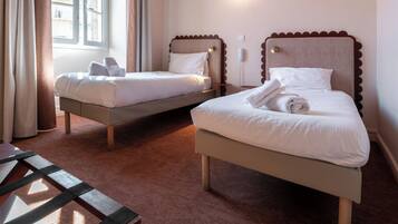 A hotel room with two beds, a wooden bench, a window with curtains, and a wall-mounted light.