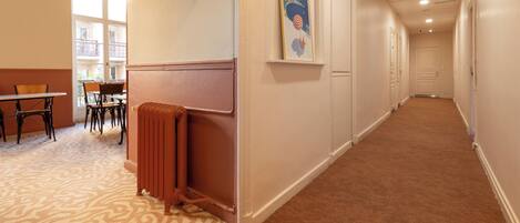 A hotel hallway with a carpeted floor, a radiator, a dining area with chairs and a table, and a poster on the wall.