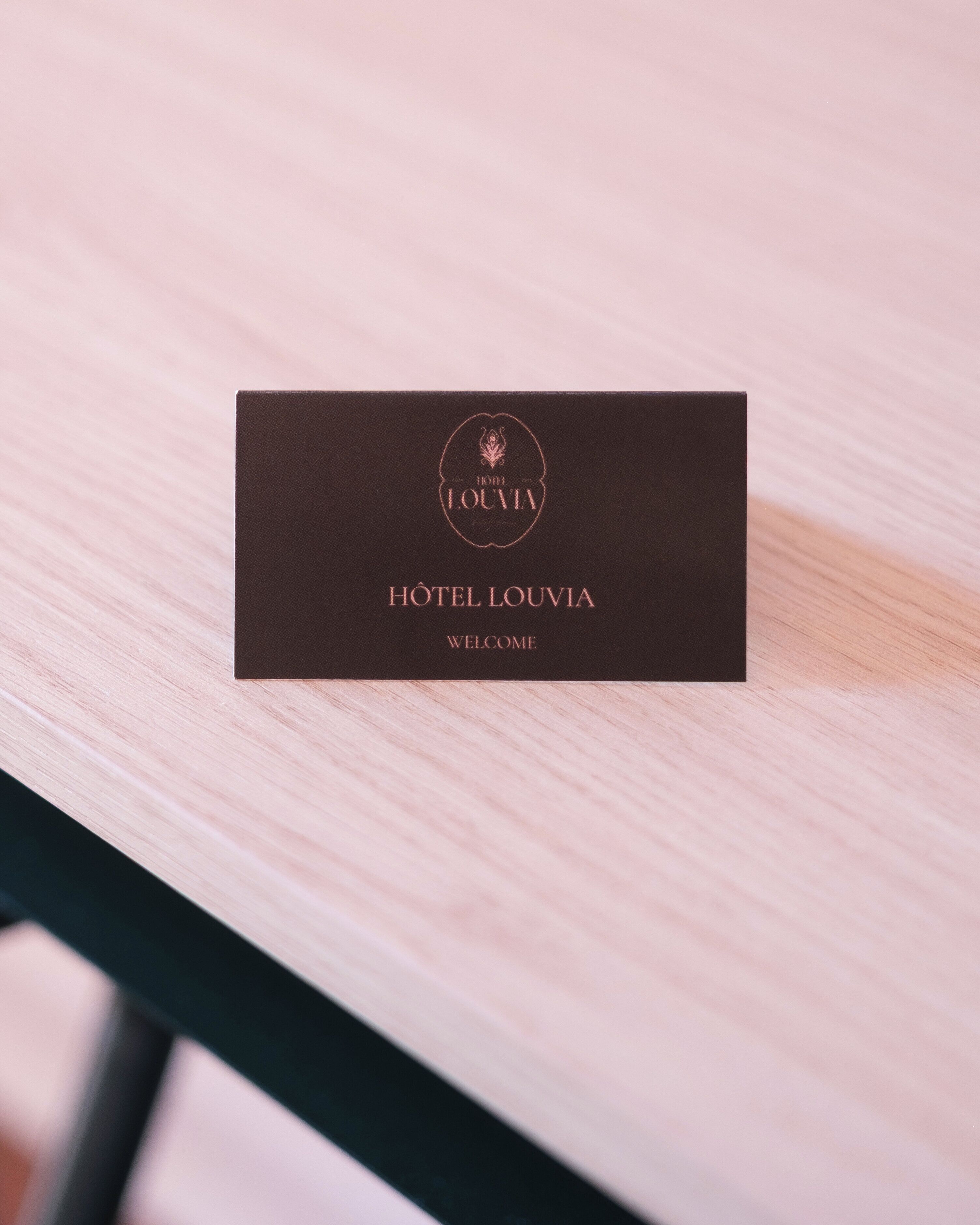 A dark brown business card for Hotel Louvia with the text 'HOTEL LOUVIA' and 'WELCOME' on a light wooden surface.