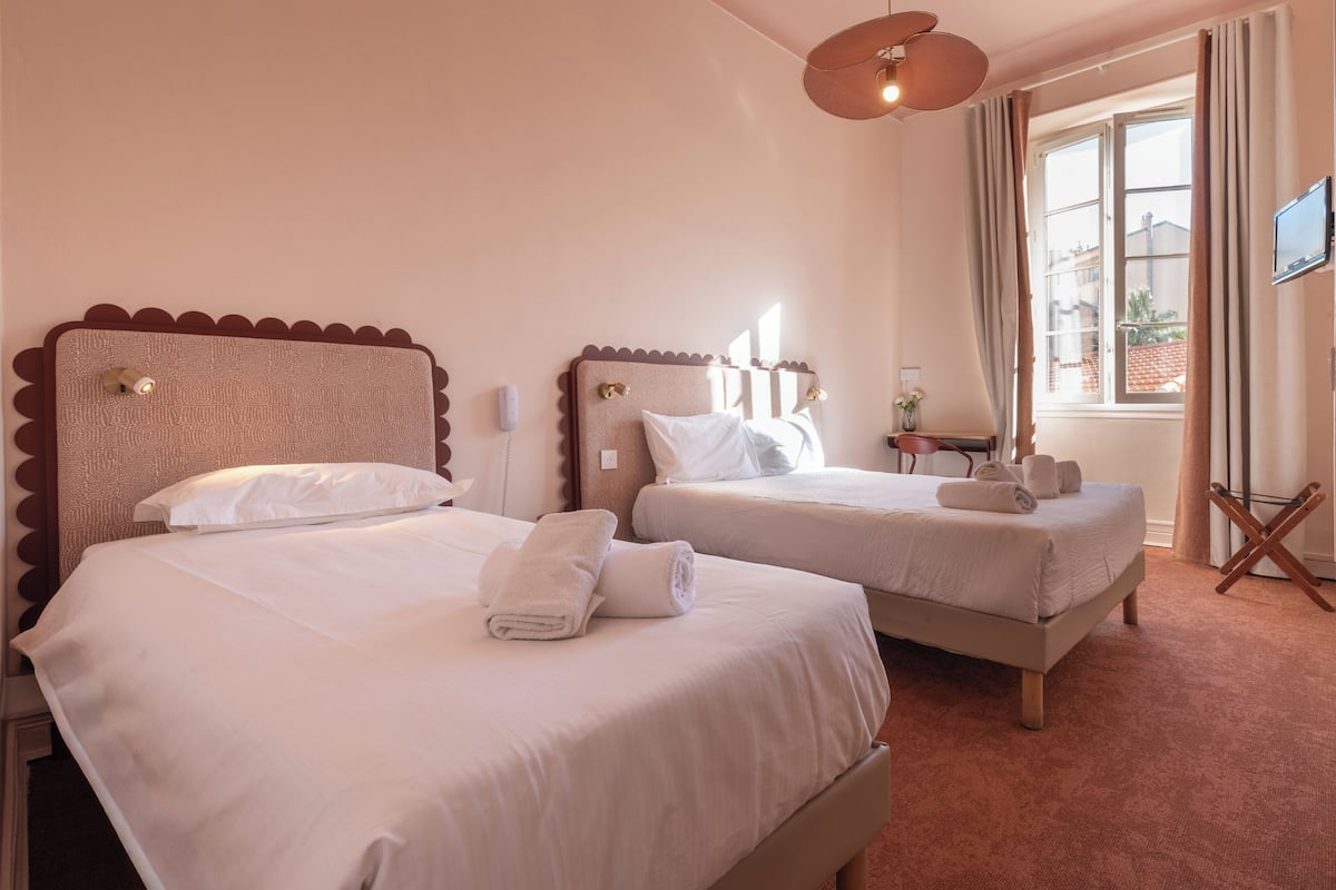 A hotel room with two beds, a flat-screen TV, a small table, and a window with curtains.