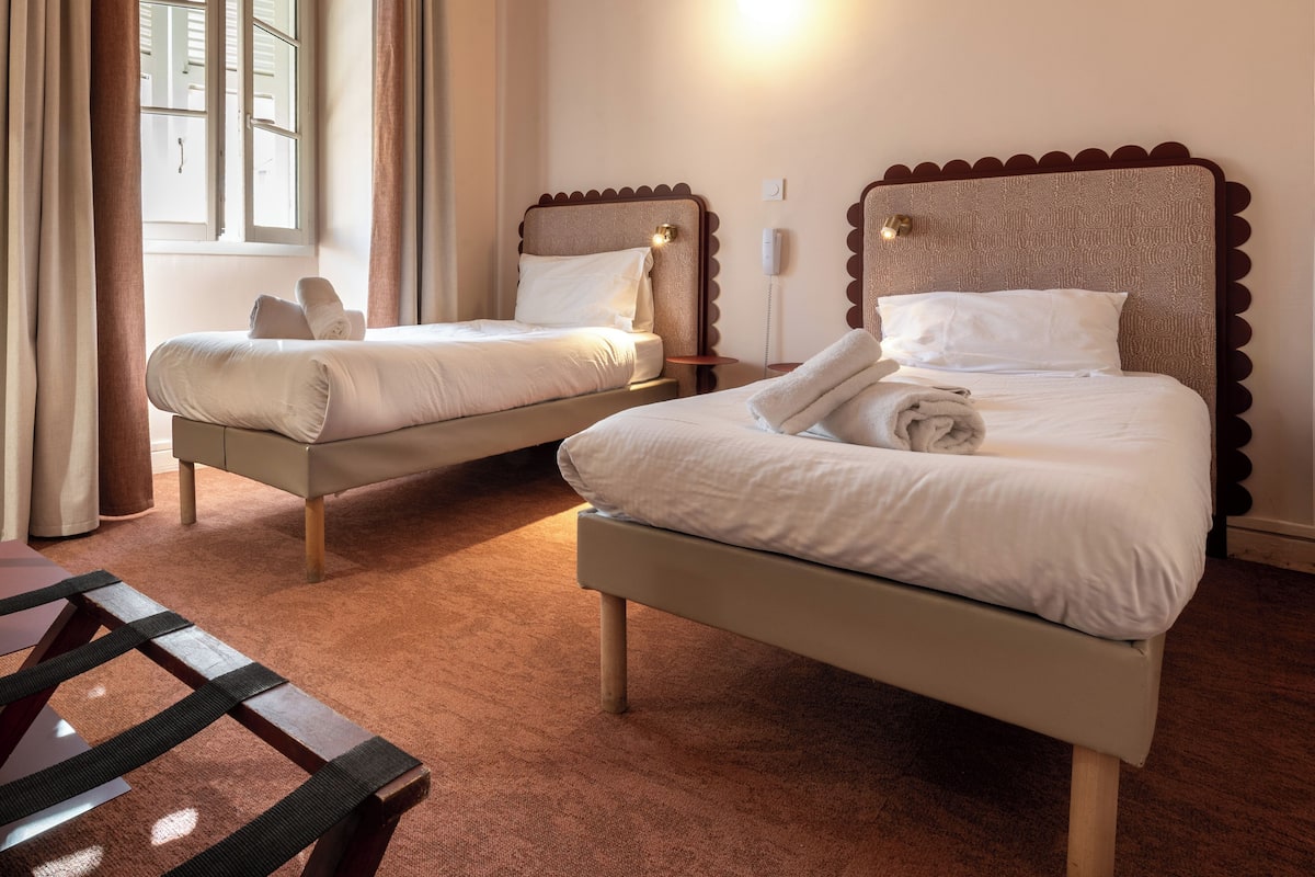 A hotel room with two beds, a wooden headboard, a bedside table, and a window with curtains.