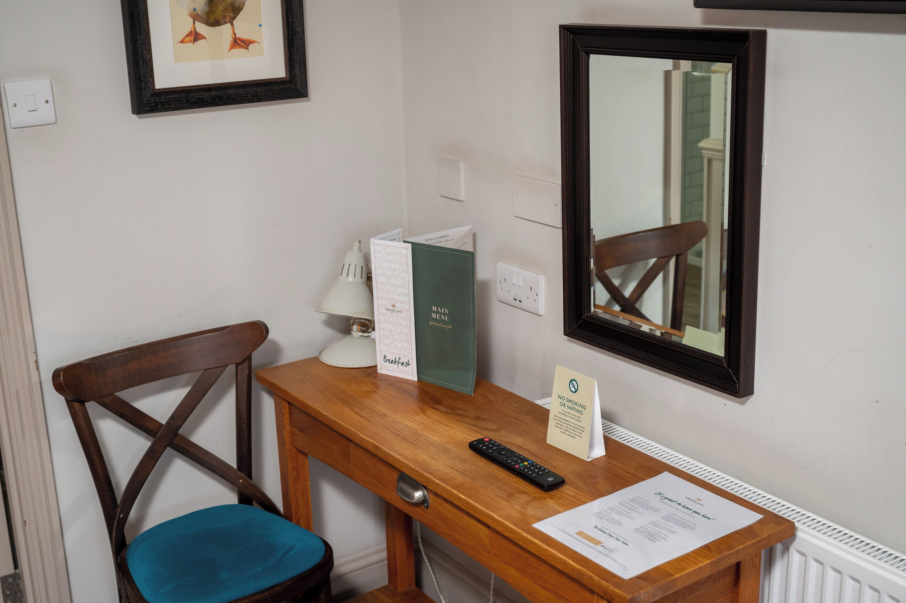Une chambre d’hôtel avec un bureau en bois, une chaise, un tableau encadré au mur, un miroir et un menu posé sur le bureau.
