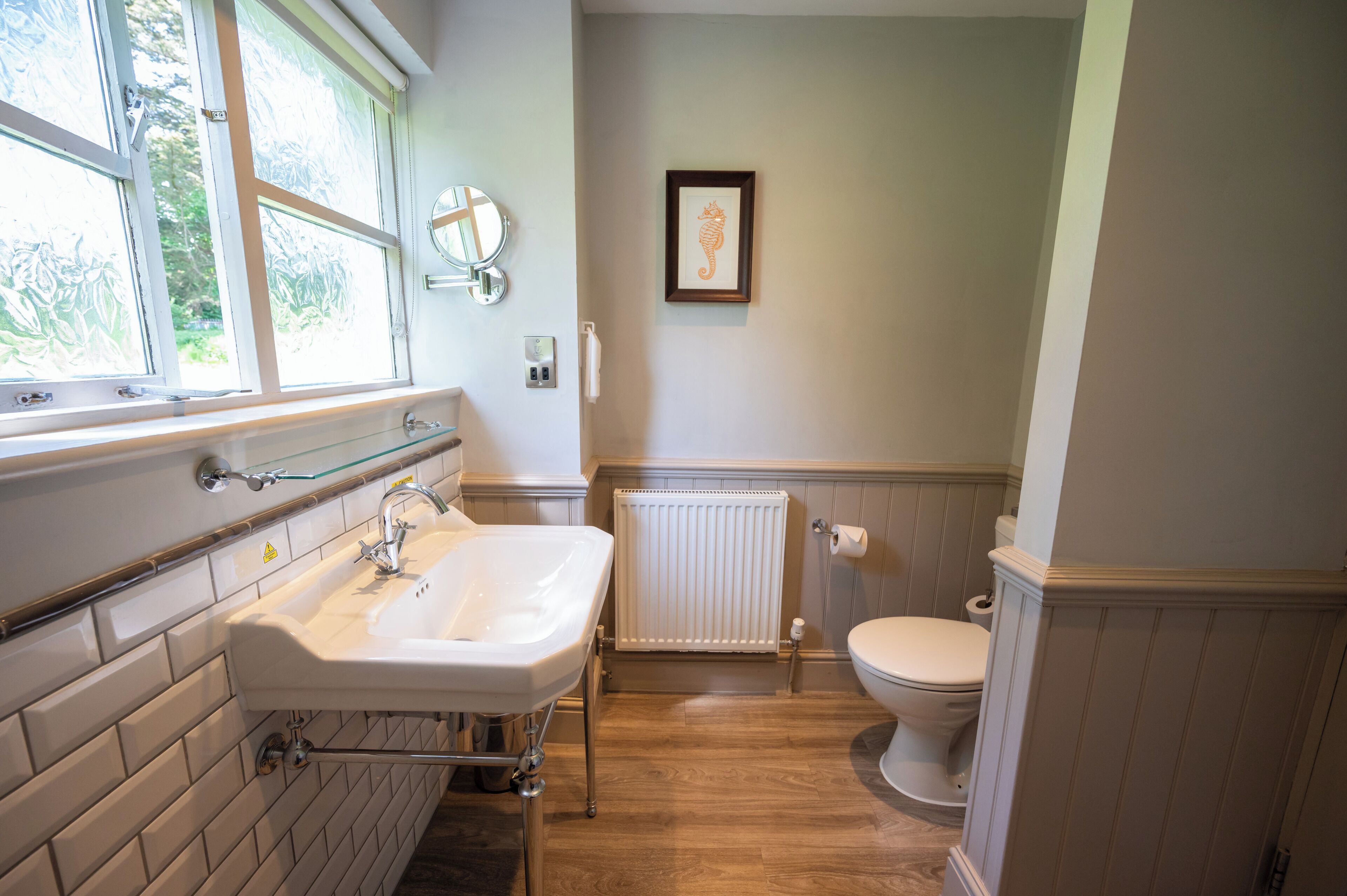 A bathroom with a white sink, a toilet, a framed seahorse artwork, and a window with a view of greenery.