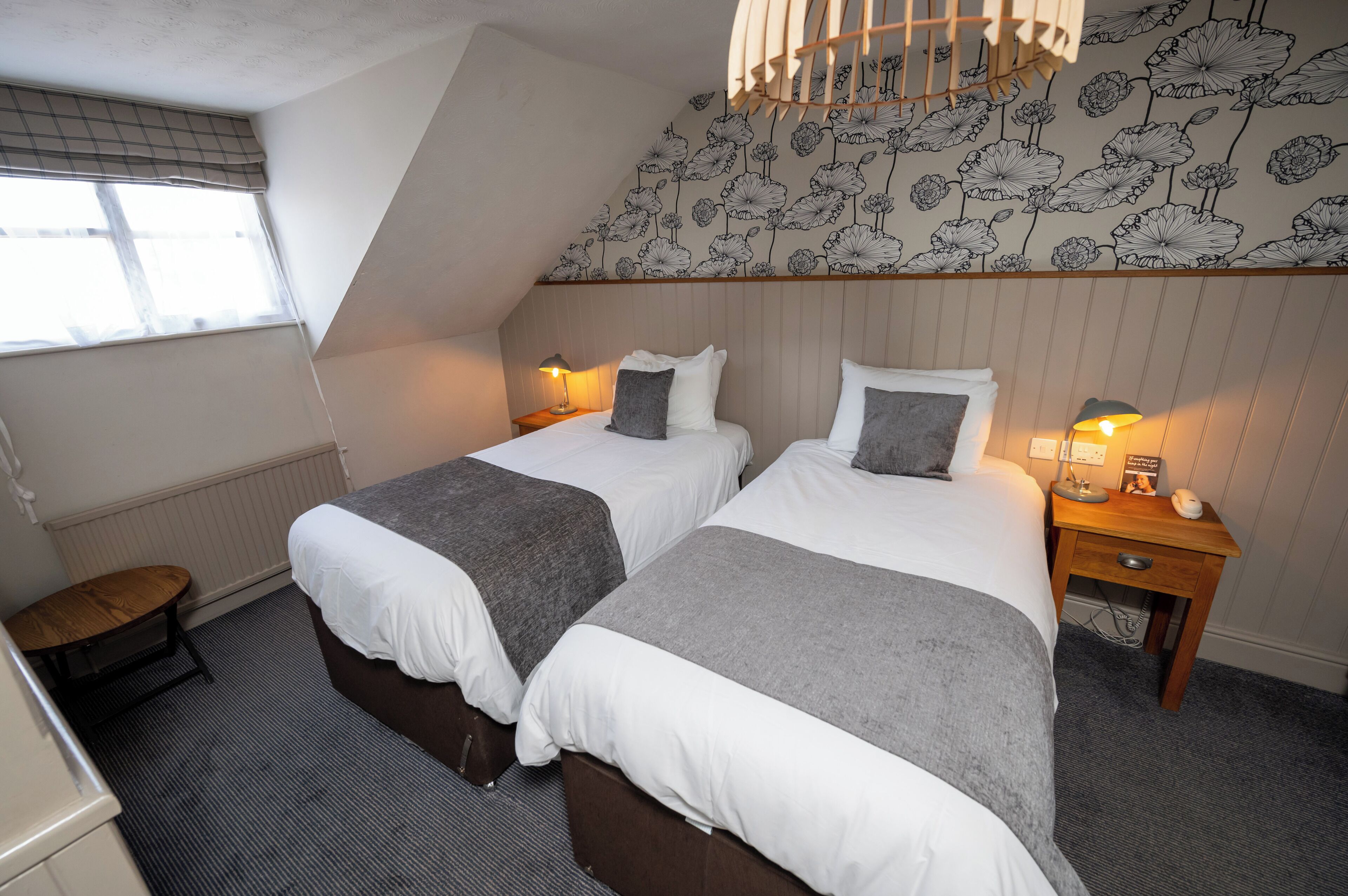 A bedroom with two beds, a window with blinds, a radiator, a small table, and a nightstand with a lamp.