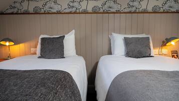 Habitación de hotel con dos camas, cada una con ropa blanca y almohadones grises. La habitación presenta un diseño de papel tapiz floral y un cabecero de madera.