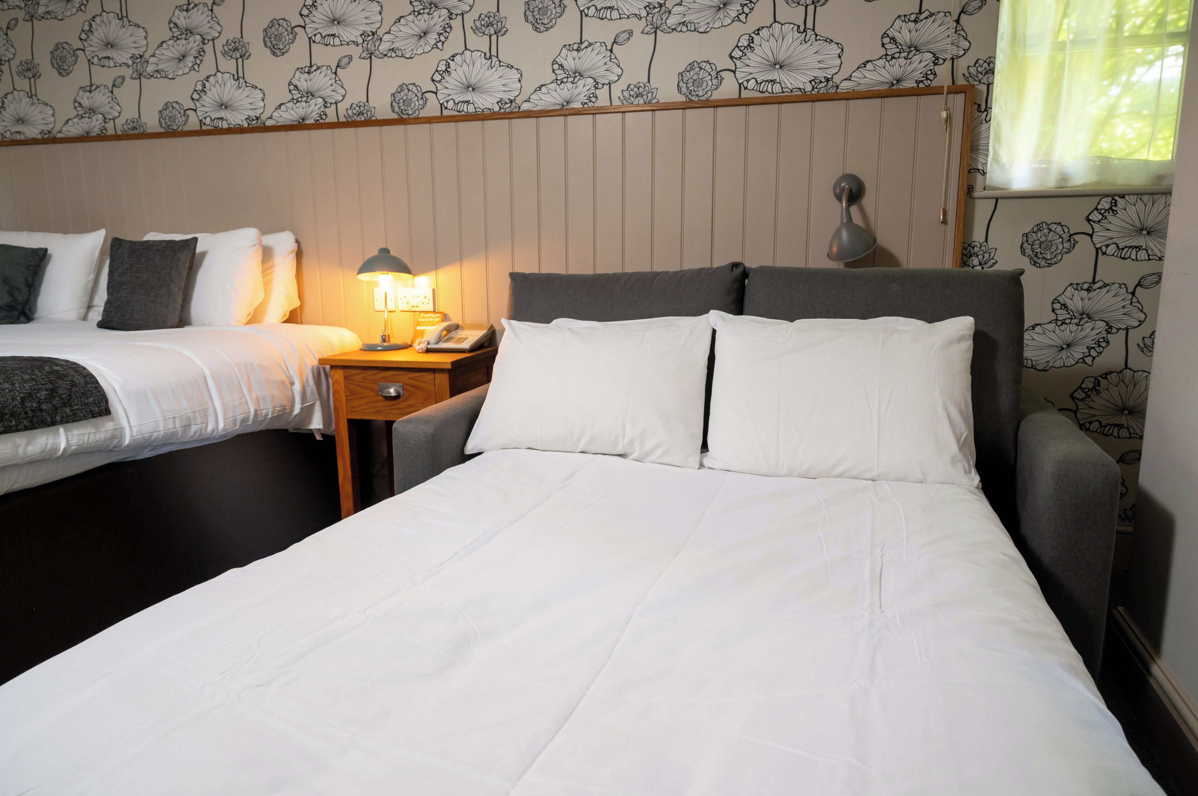A hotel room with two beds, a nightstand with a lamp, and floral wallpaper.