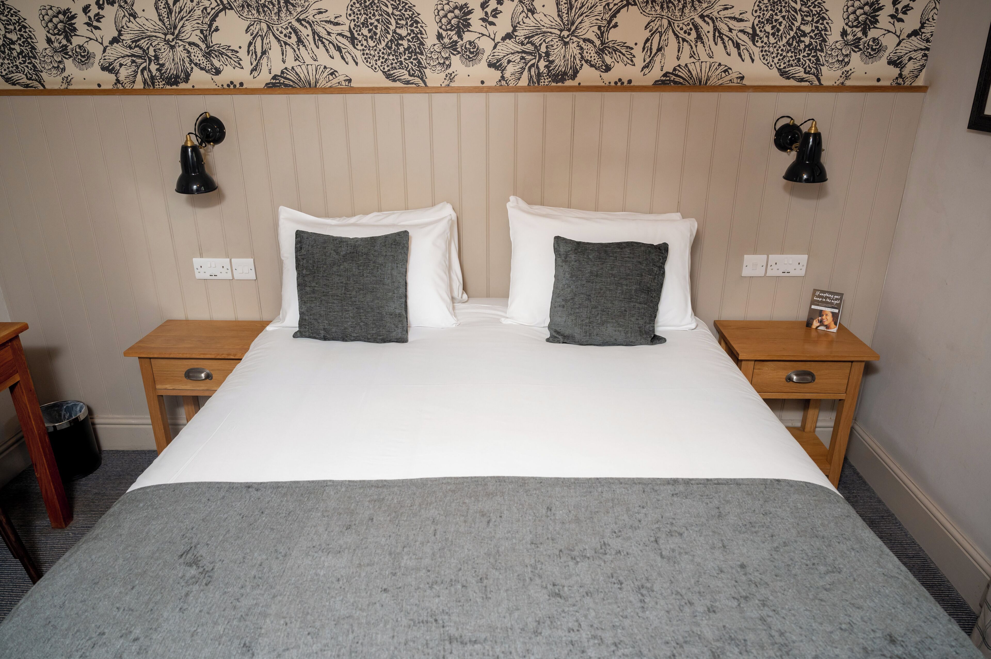 A neatly made bed with two pillows, a bedside table with a lamp, and a wall with floral wallpaper.