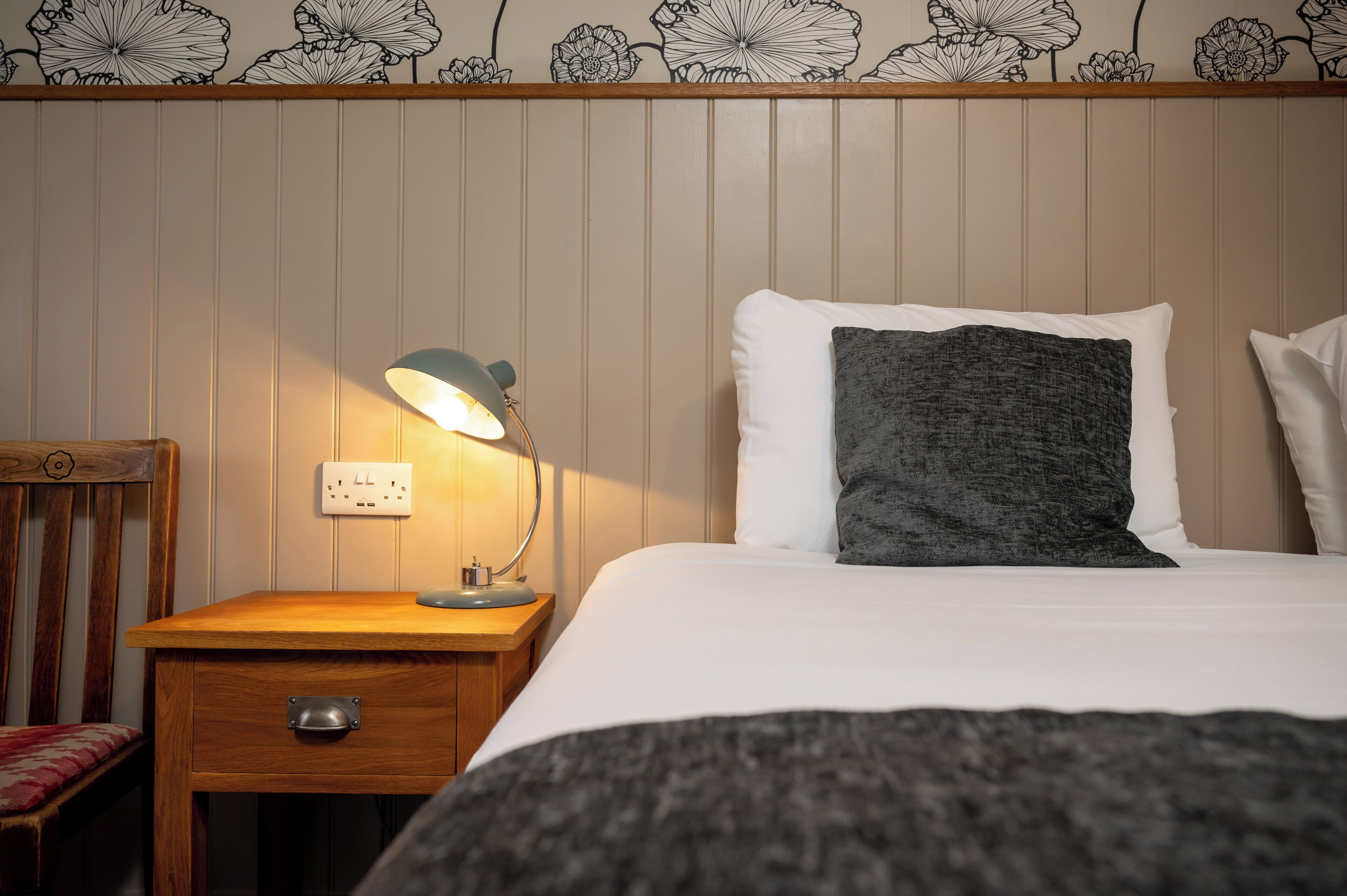 A neatly made bed with white and gray pillows, a wooden nightstand with a lamp, and a chair with a red cushion.