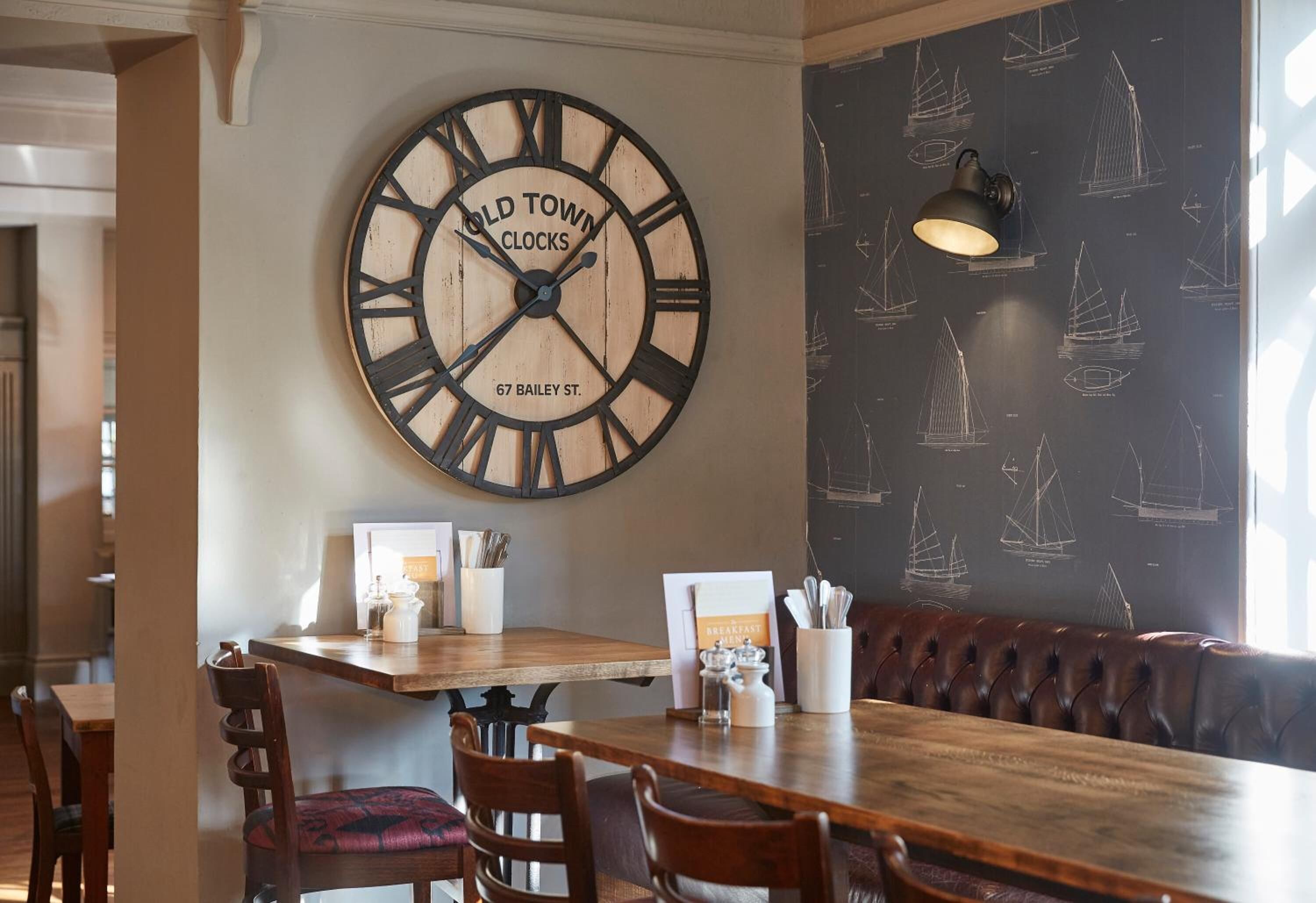Interior de restaurante con un gran reloj de pared, mesas de madera, asientos acolchados y un papel tapiz con motivos náuticos.