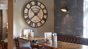 Interior de restaurante con un gran reloj de pared, mesas de madera, asientos acolchados y un papel tapiz con motivos náuticos.