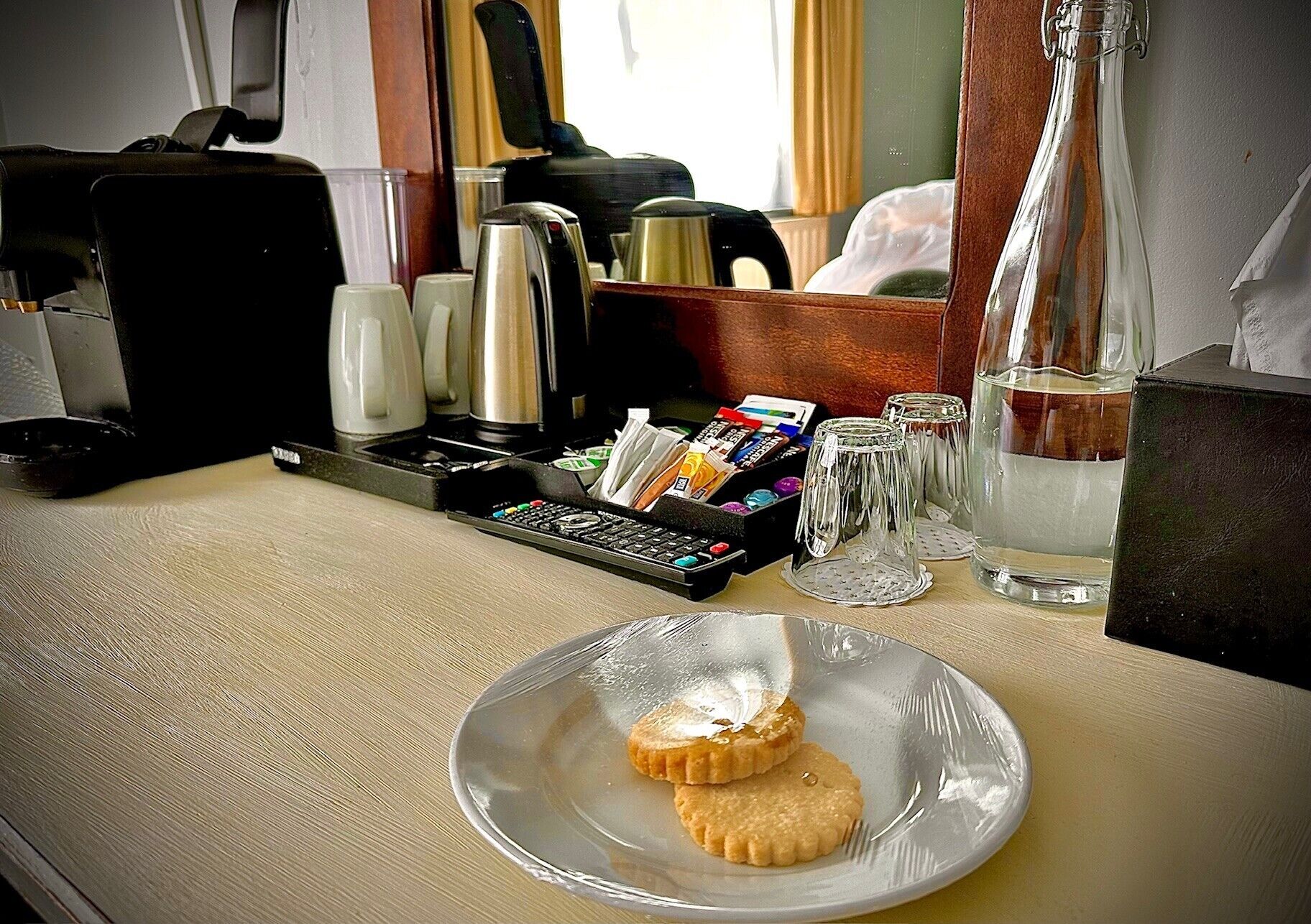 A breakfast table with a plate of biscuits, a coffee maker, a kettle, a glass of water, and a small box of tea bags.