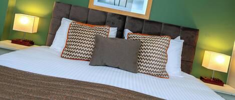 A neatly made bed with patterned pillows, a framed picture above, and bedside lamps on green walls.