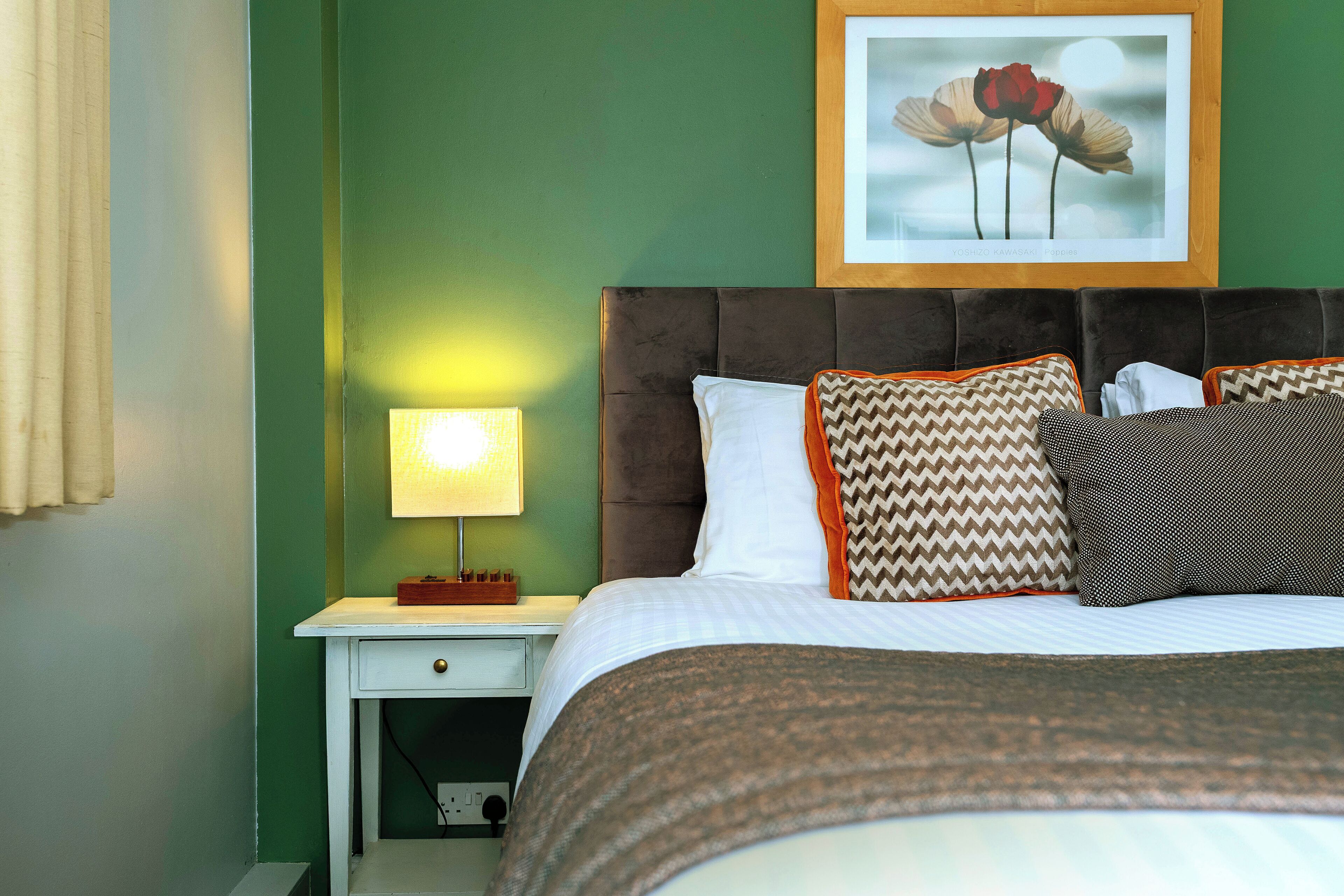 Un letto ben rifatto con cuscini decorati, un quadro appeso sopra e una lampada da comodino su un comodino bianco.