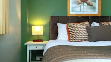 Un letto ben rifatto con cuscini decorati, un quadro appeso sopra e una lampada da comodino su un comodino bianco.