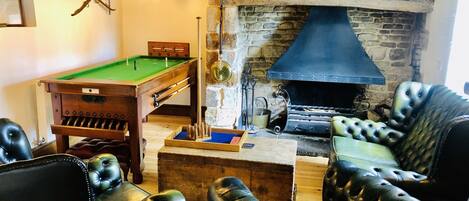 A billiards table, a fireplace, and leather armchairs in a room with exposed brick walls and wooden beams.
