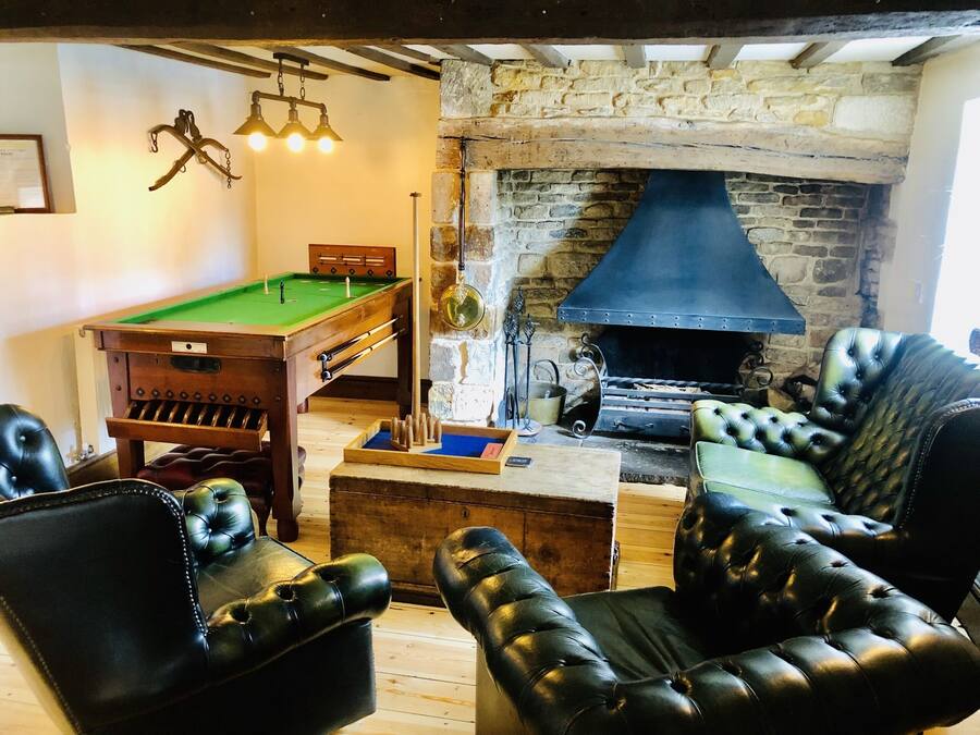 A billiards table, a fireplace, and leather armchairs in a room with exposed brick walls and wooden beams.