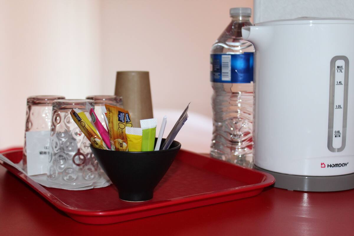 Una bandeja roja con una taza negra que contiene bolígrafos y una botella de agua, una cafetera blanca y un recipiente de vidrio con una pajita.