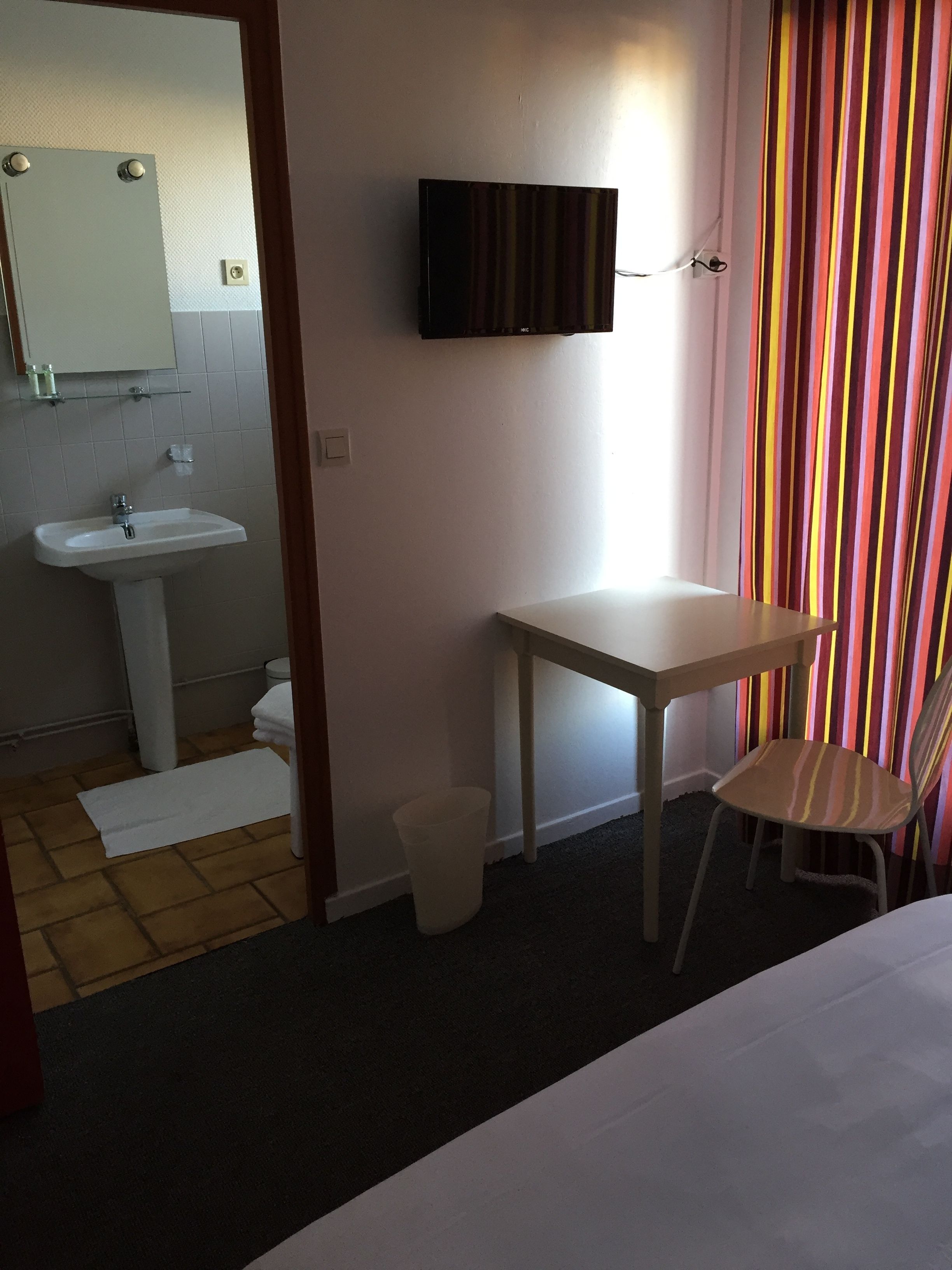 Habitación de hotel con cama, una mesita, una silla, televisor montado en la pared, lavamanos y espejo.