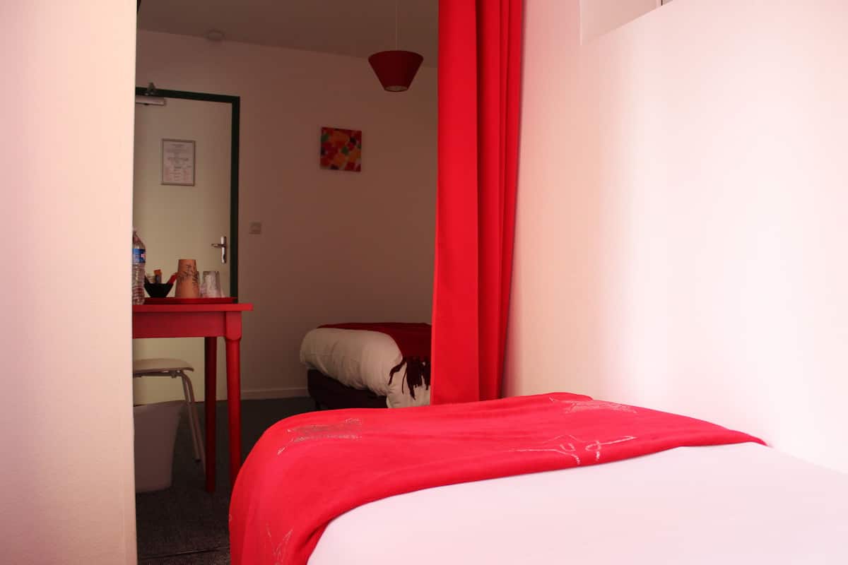 Un dormitorio con cama, una cortina roja, un escritorio con silla y una mesita con una botella y una taza.