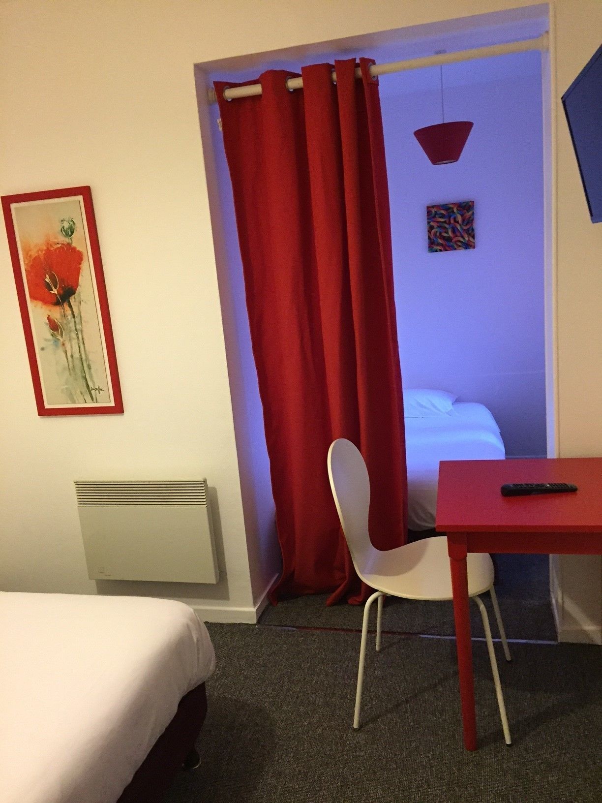 Una habitación de hotel con cama, escritorio, silla, cortina roja, radiador y un cuadro en la pared.