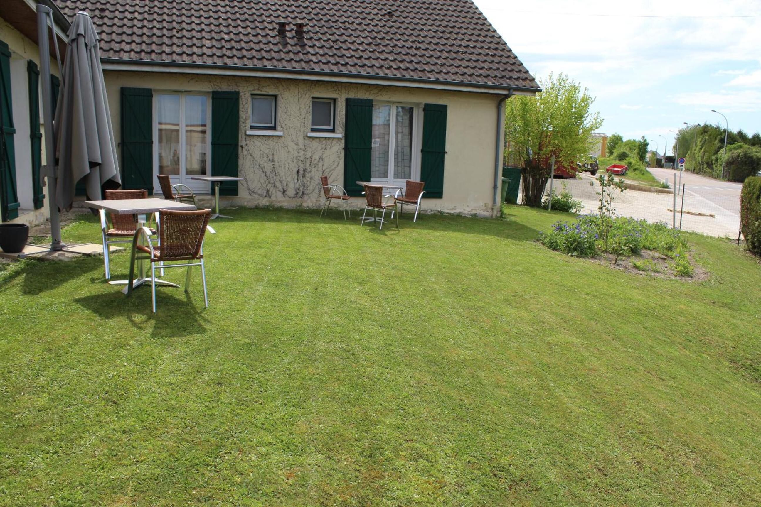A house with a tiled roof, green shutters, and a well-maintained lawn. There are two outdoor tables and chairs.