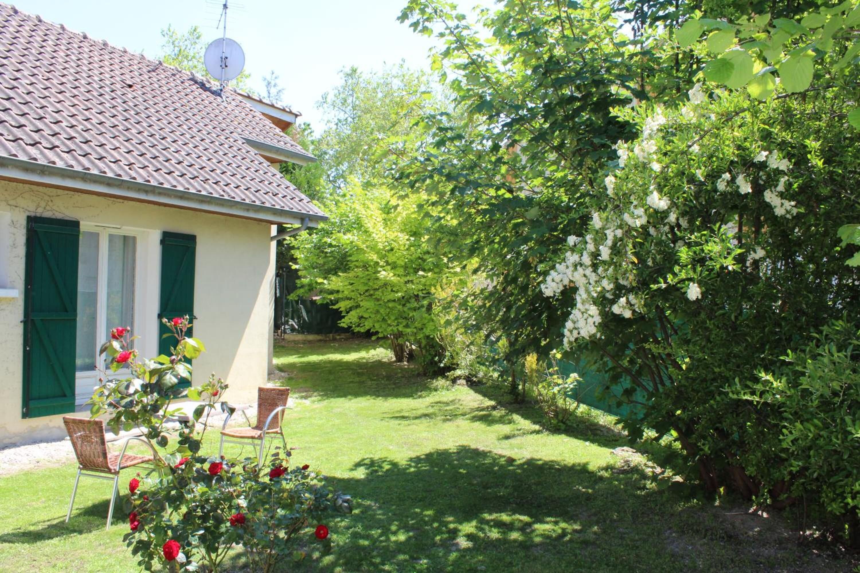 A house with a tiled roof, green shutters, and a garden with a lawn and flowering bushes.