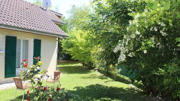A house with a tiled roof, green shutters, and a garden with a lawn and flowering bushes.