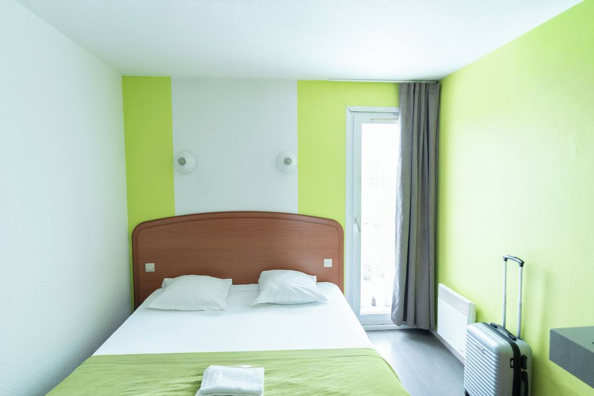 Ein Hotelzimmer mit Bett, Heizkörper, einem Fenster mit Vorhängen und einem Koffer.