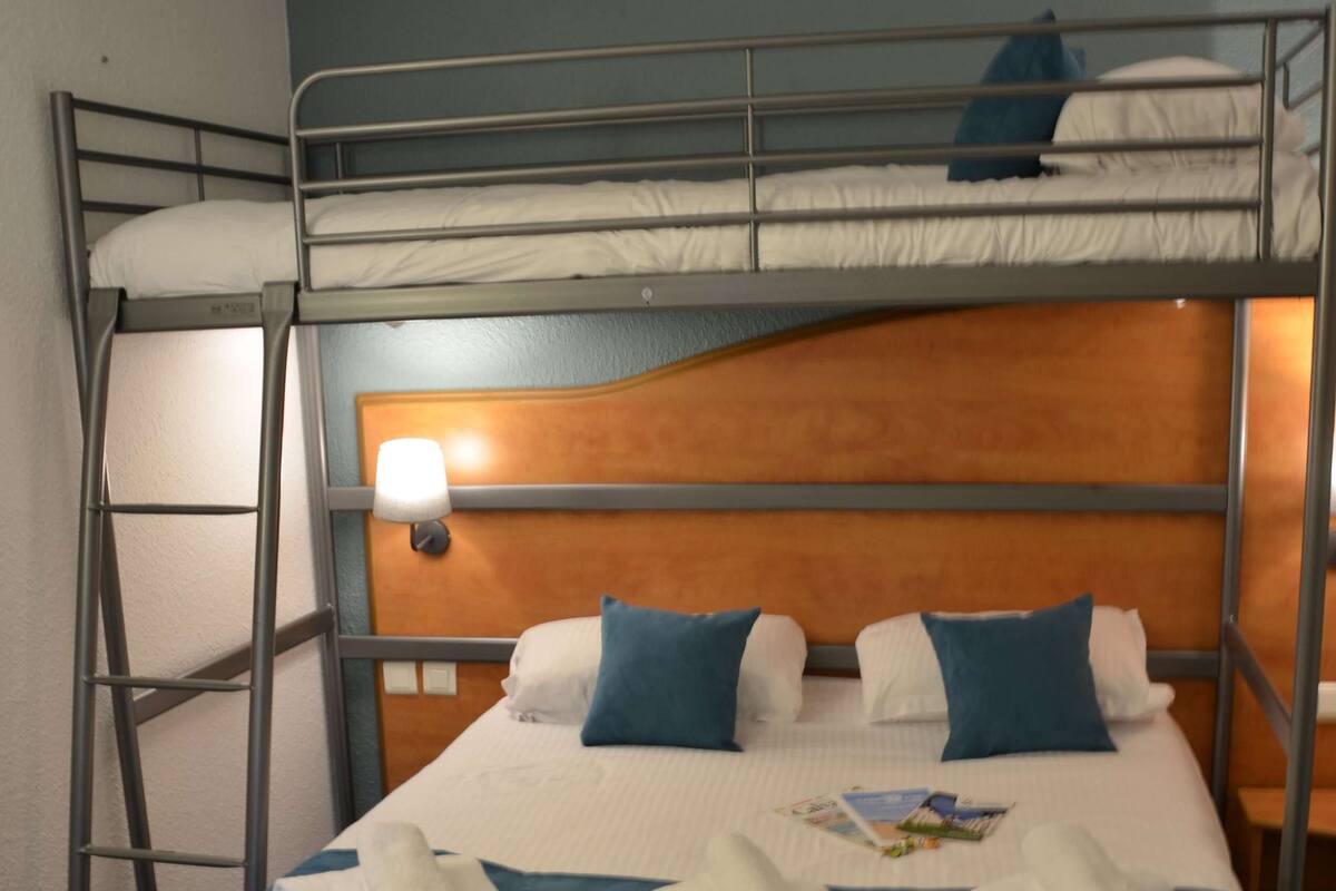 Un lit superposé avec une tête de lit en bois, une table de chevet avec une lampe et des serviettes soigneusement pliées sur le lit du bas.