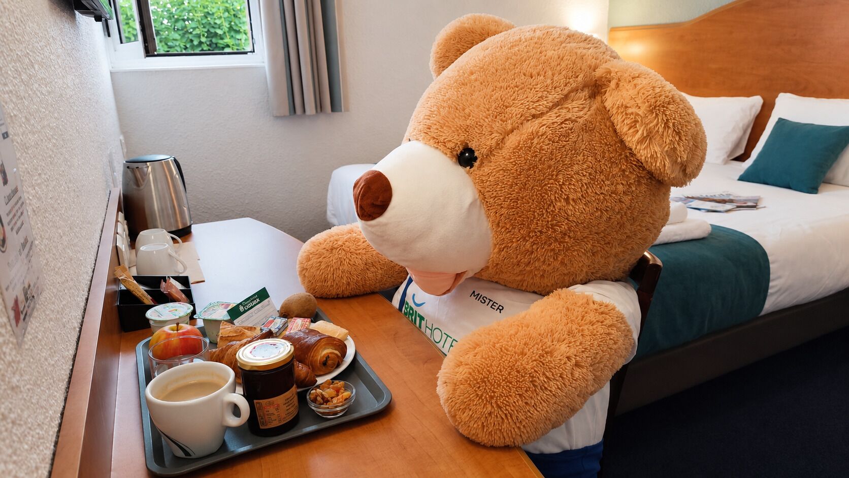 Un osito de peluche en una habitación de hotel, con una bandeja de alimentos para el desayuno y una cafetera.
