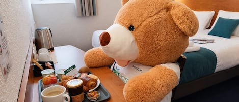 Un osito de peluche en una habitación de hotel, con una bandeja de alimentos para el desayuno y una cafetera.