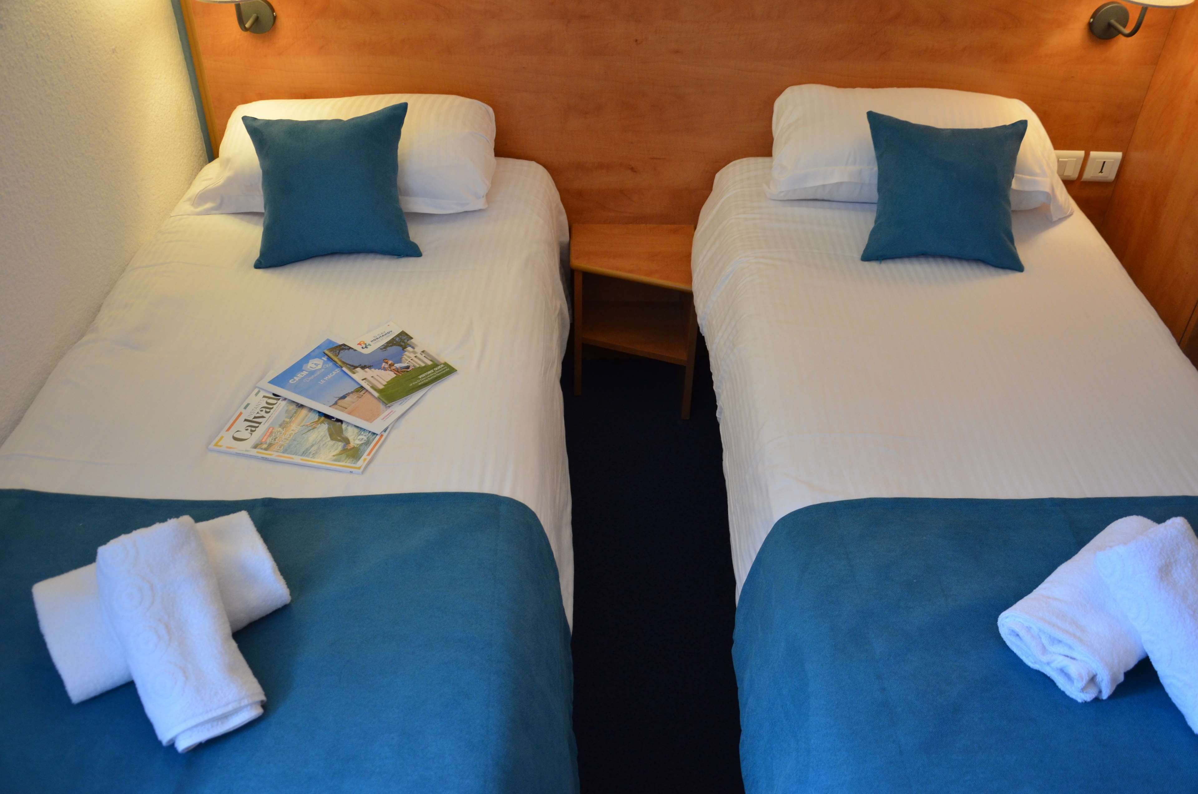 Two single beds with blue bedding and white pillows, a folded white towel on each bed, and a magazine on the left bed.