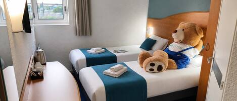 Una habitación de hotel con dos camas, un escritorio, un osito de peluche y una ventana con cortinas.