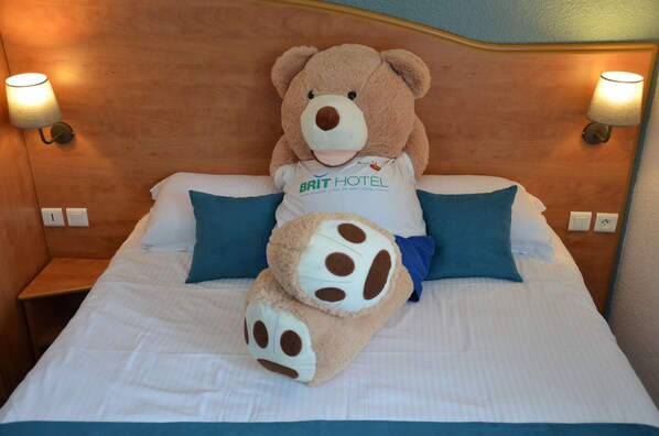 Una cama con un oso de peluche grande y un animal de peluche más pequeño, ambos con diseño de oso, sobre una colcha blanca y almohadas azules.