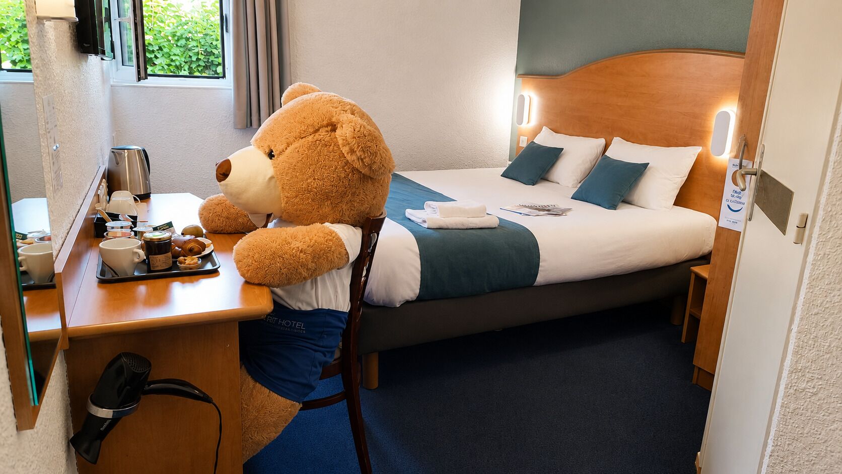 Une chambre d’hôtel avec un lit, un bureau, un grand ours en peluche et une fenêtre avec des rideaux.