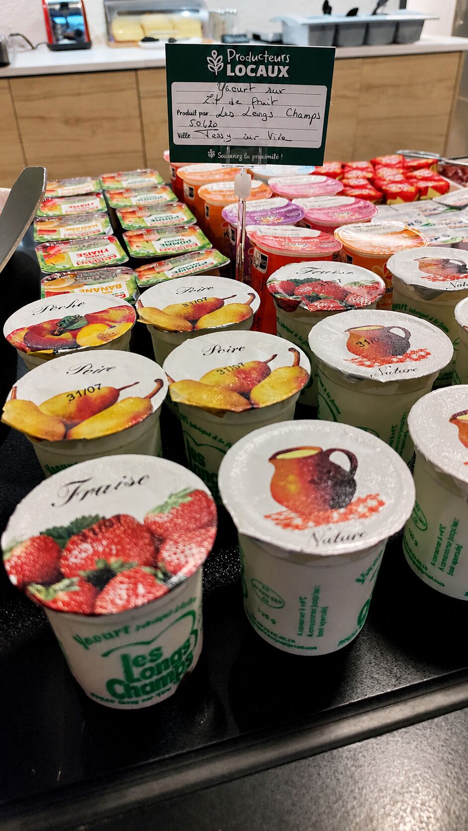 Exhibición de vasos de yogur con motivos de frutas, un letrero que indica 'Producteurs LOCAUX' y una nota sobre 'Yogurt sur le fruit'.