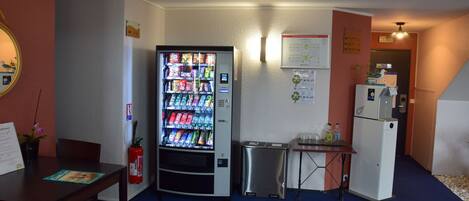 A compact hotel room with a minibar, a small table, a seating area, and a vending machine.