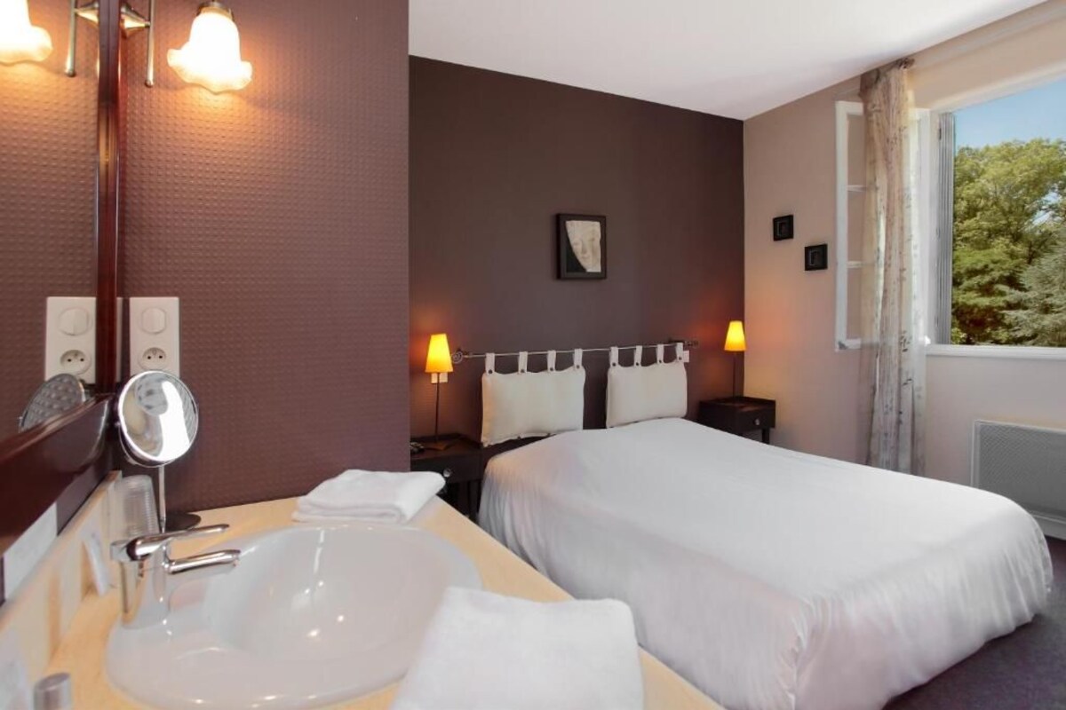 A hotel room with a large bed, a bedside table with lamps, a mirror, and a view of trees through the window.
