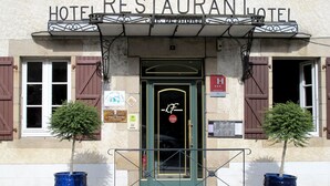 A hotel with a sign reading "HOTEL RESTAURANT HOTEL DE SHOR". There are two potted trees in front of the entrance, and a sign indicating Michelin star rating. - Deshors Foujanet (Chamboulive)