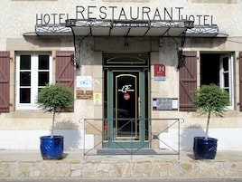 Um hotel com uma placa que diz 'HOTEL RESTAURANT HOTEL DE SHOR'. Há duas árvores em vasos na frente da entrada, e uma placa indicando classificação Michelin.