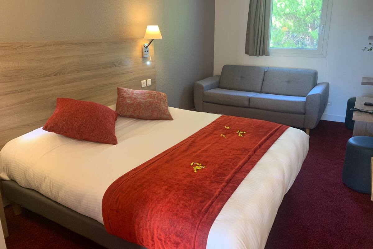 Une chambre d’hôtel avec un lit, un canapé gris, une fenêtre avec des rideaux et une lampe fixée au mur.