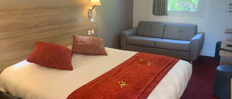 Una habitación de hotel con una cama, un sofá gris, una ventana con cortinas y una lámpara fijada a la pared.