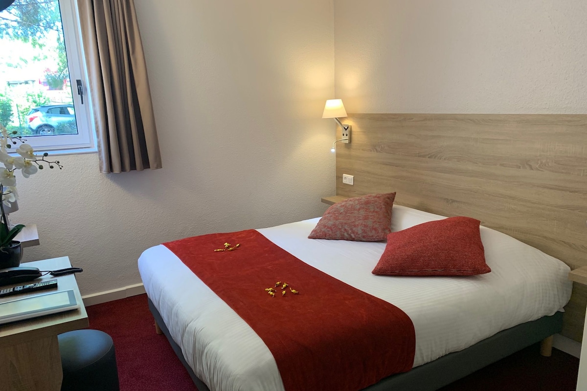 A hotel room with a bed, a desk with a phone, a lamp, and a window with curtains.