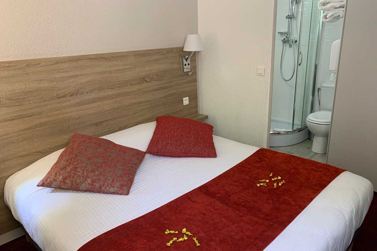 A hotel room with a wooden headboard, a bed with white bedding and red pillows, a red throw blanket with gold accents, a white lamp, and a glass-enclosed shower area.