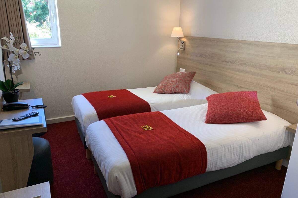 Une chambre d’hôtel avec deux lits, chacun recouvert d’une couverture rouge et muni d’un oreiller. On y trouve un bureau avec un téléphone, une télécommande et une lampe. Une fenêtre avec des rideaux et une plante est visible.