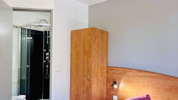 A bedroom with a wooden wardrobe, a bed with a patterned pillow, and a bathroom with a glass shower enclosure.