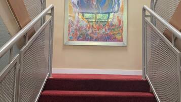 Una escalera con peldaños cubiertos de moqueta roja y barandillas de metal, que conduce a una pared con un cuadro abstracto y colorido.