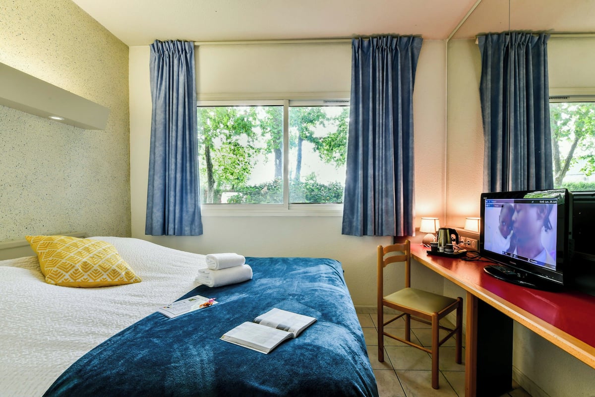 Una habitación de hotel con cama, escritorio con televisor, silla y ventana con cortinas.
