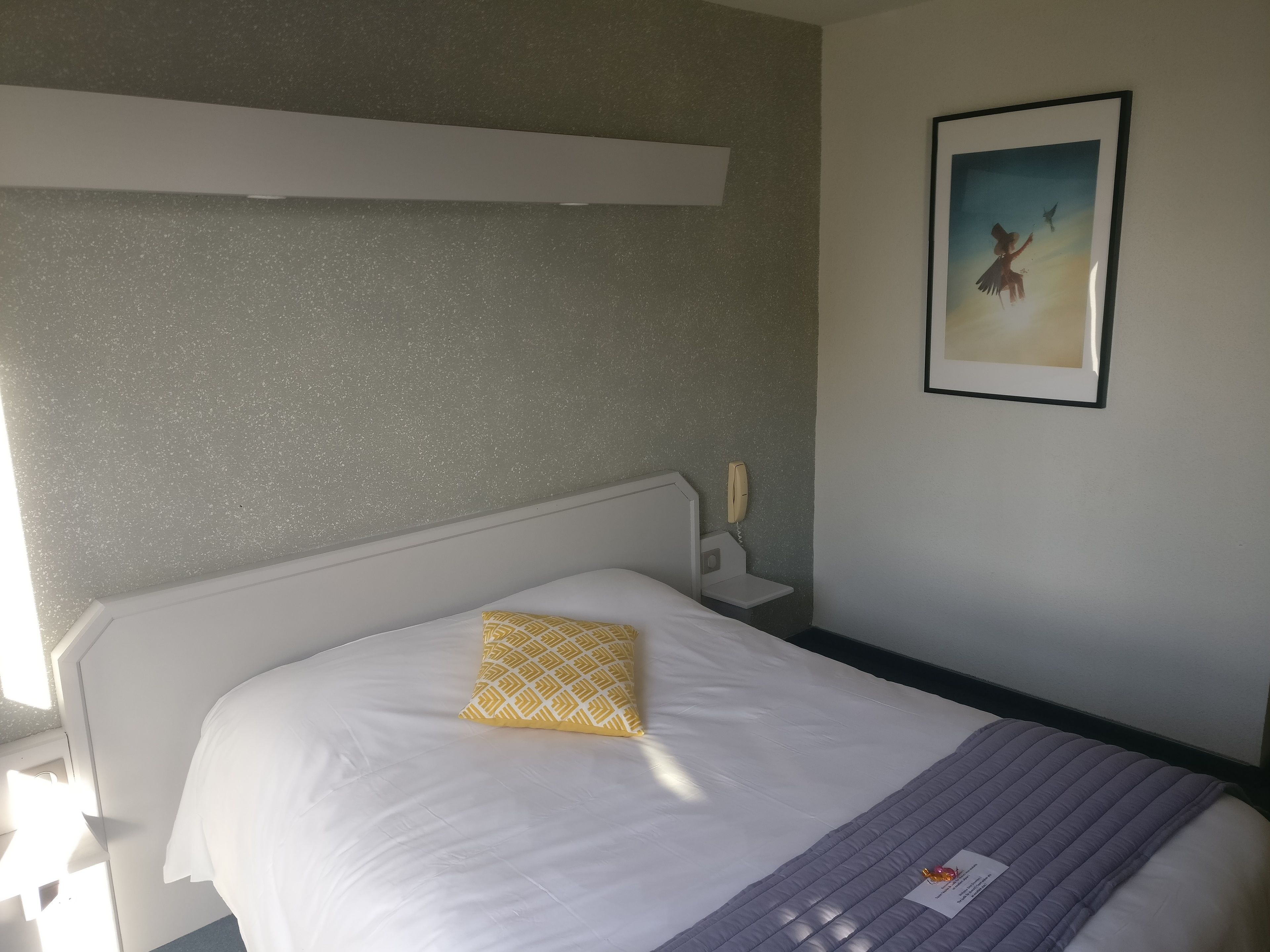 A hotel room with a bed, a bedside table, a telephone, and a framed picture on the wall.